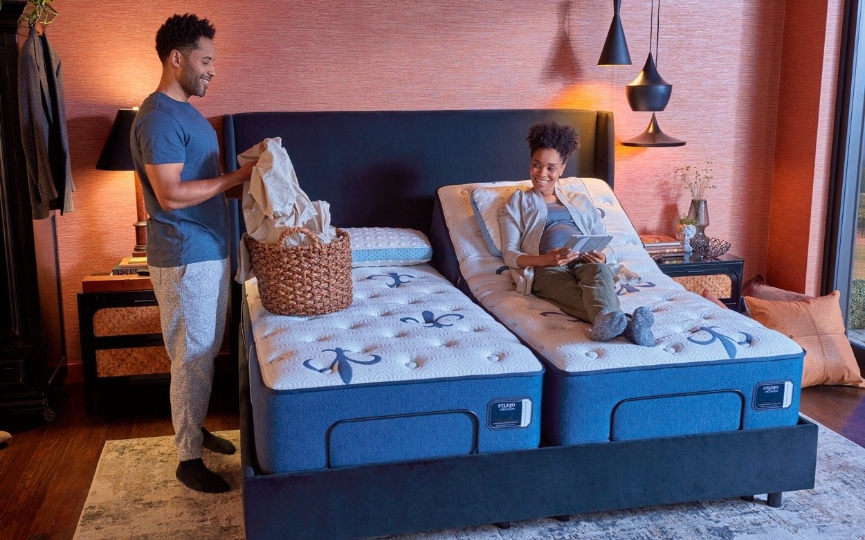 Split king Studio tight top mattress, on an adjustable base, in a dressed room, with couple lounging. One side of bed's head is raise.